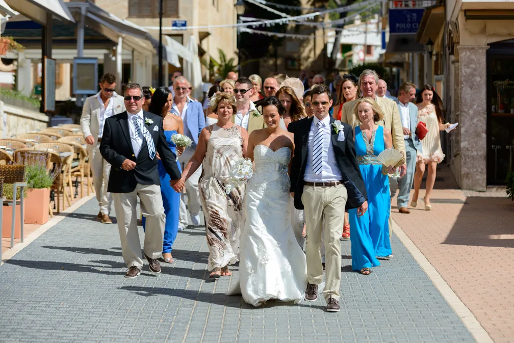 Bride in white wedding dress walking arm-in-arm with groom and guests on a sunny paved street.