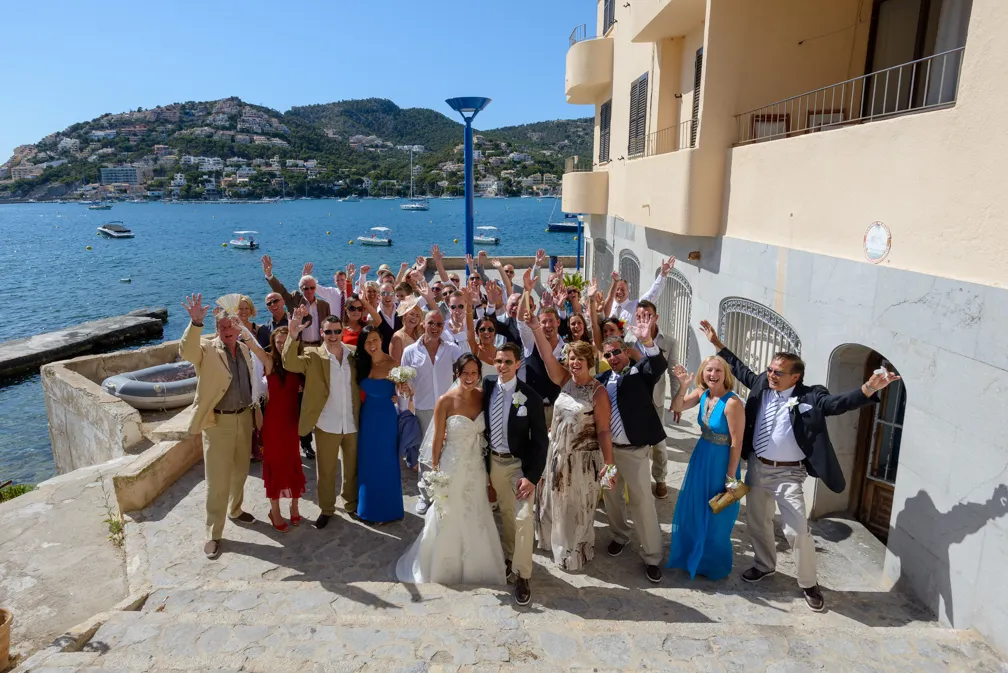 Group of wedding guests and the bride and groom posing cheerfully on a stone pathway by the water with boats and hills in the background.