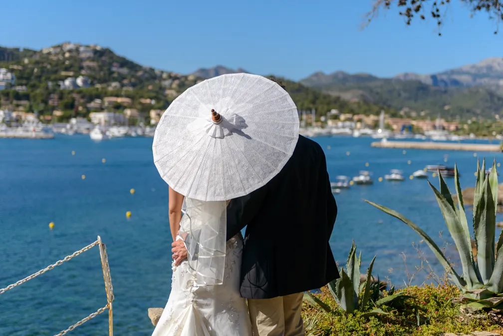 Couple embracing by a waterfront with the bride holding a white lace parasol, overlooking boats and hills on a sunny day.