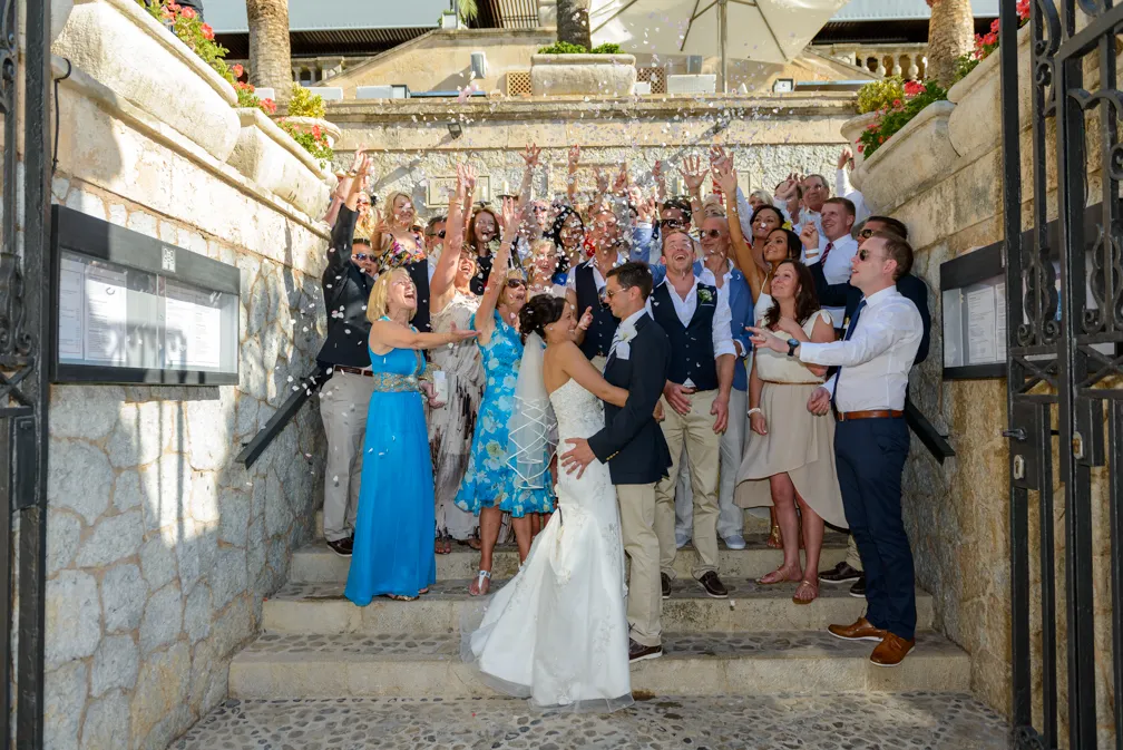 Bride and groom embracing on steps surrounded by cheering guests tossing flower petals.