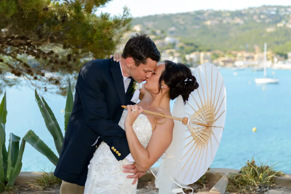 Bride and groom kissing outdoors near water, with bride holding a white parasol and greenery around.