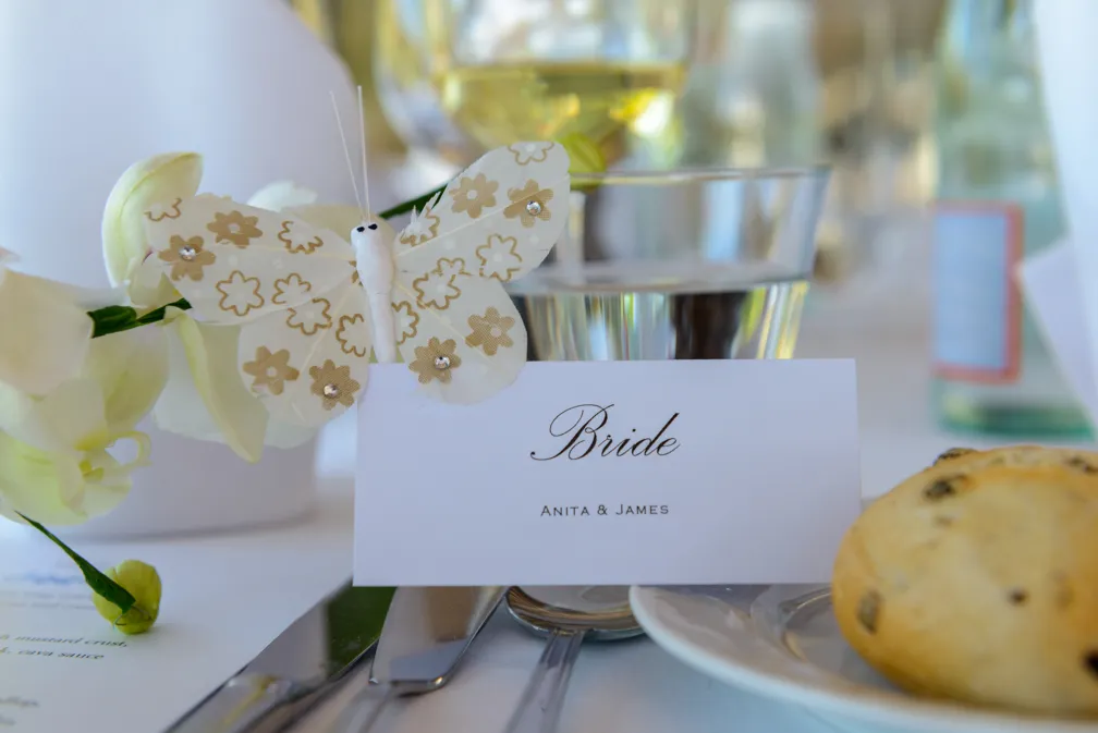 Wedding place card with 'Bride Anita & James' in front of a decorative butterfly, next to a glass, silverware, and bread roll.