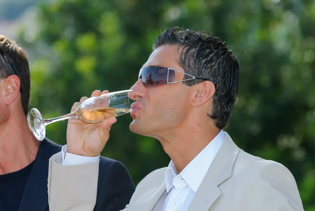 Man wearing sunglasses and a light-colored suit drinking champagne from a flute glass outdoors.