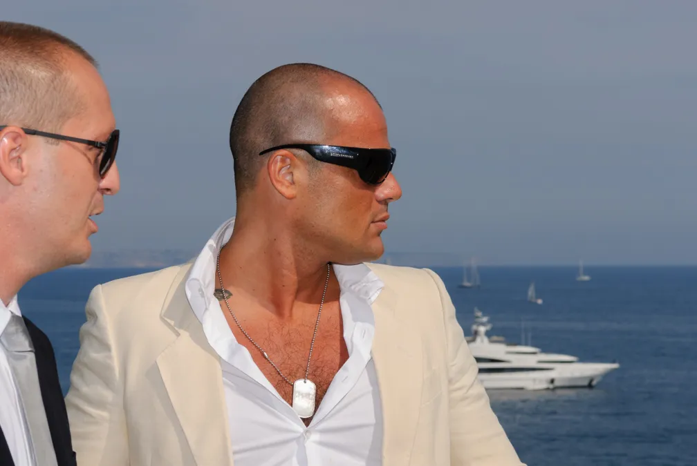 Two men wearing sunglasses, one in a white suit jacket and open-collared shirt, with a yacht and sailboats in the ocean behind them.