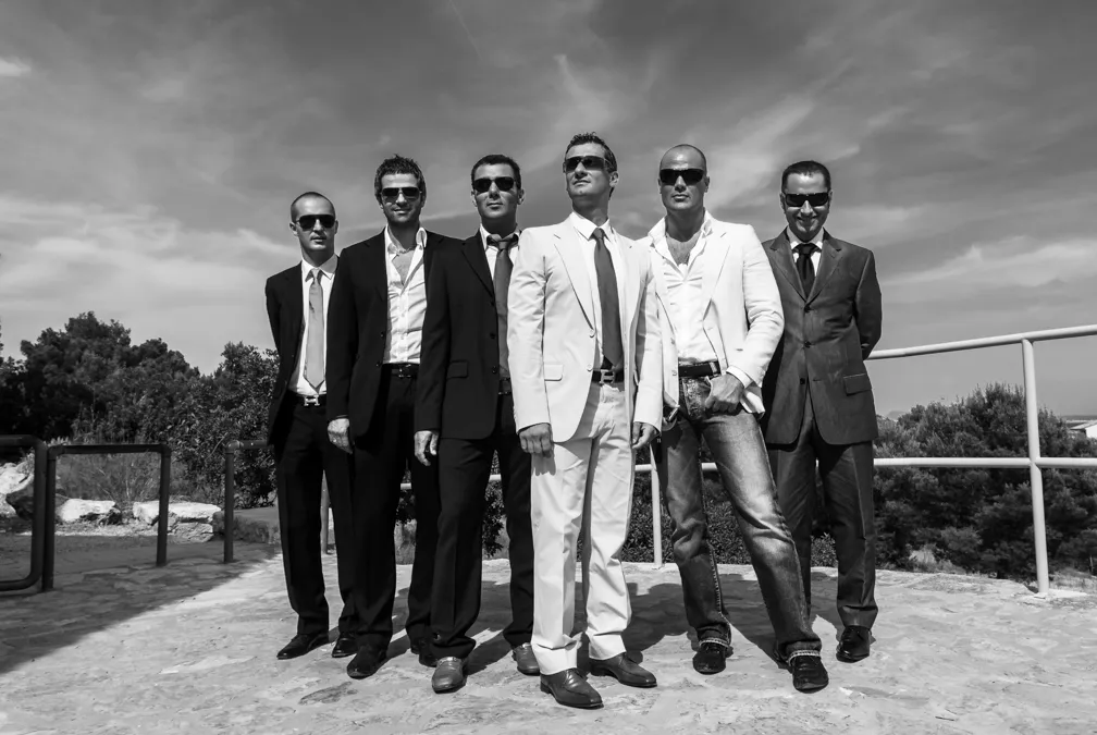 Six men wearing suits and sunglasses standing outdoors on stone ground with trees and cloudy sky in the background.