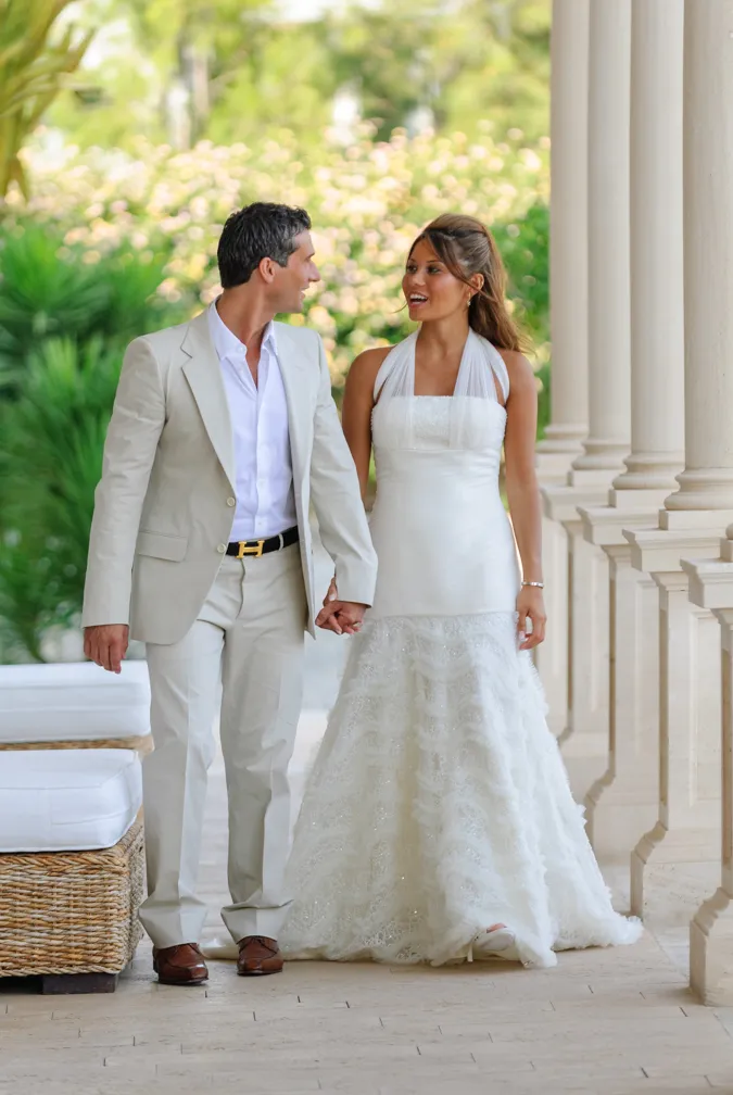 Bride in white wedding dress and groom in light beige suit holding hands and walking along a colonnade with greenery in the background.