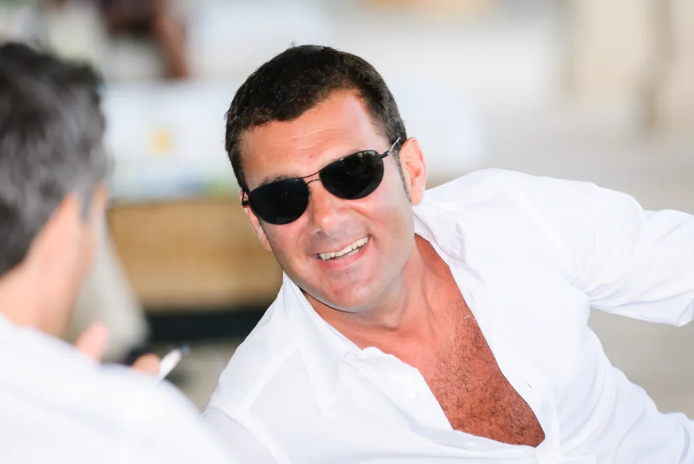 Smiling man wearing dark sunglasses and a white open-collar shirt, engaged in conversation indoors.
