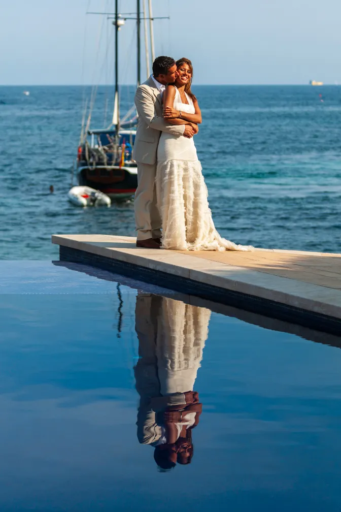 Couple embracing on a poolside pier with a sailboat and ocean in the background, their reflection visible in the water.
