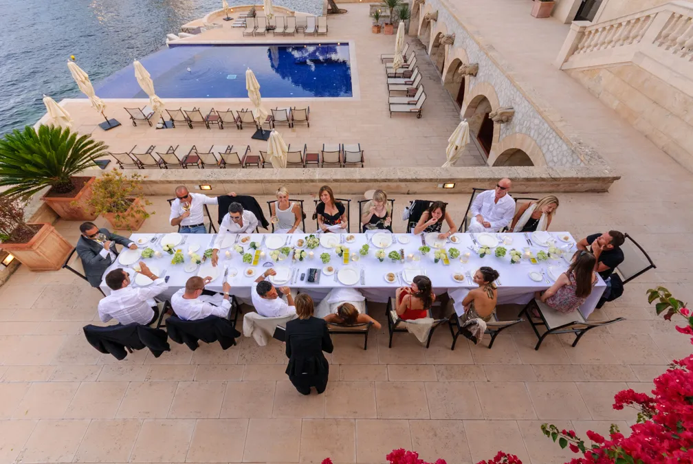 Group of people having an outdoor dinner at a long table with white tablecloth near a pool and waterfront.