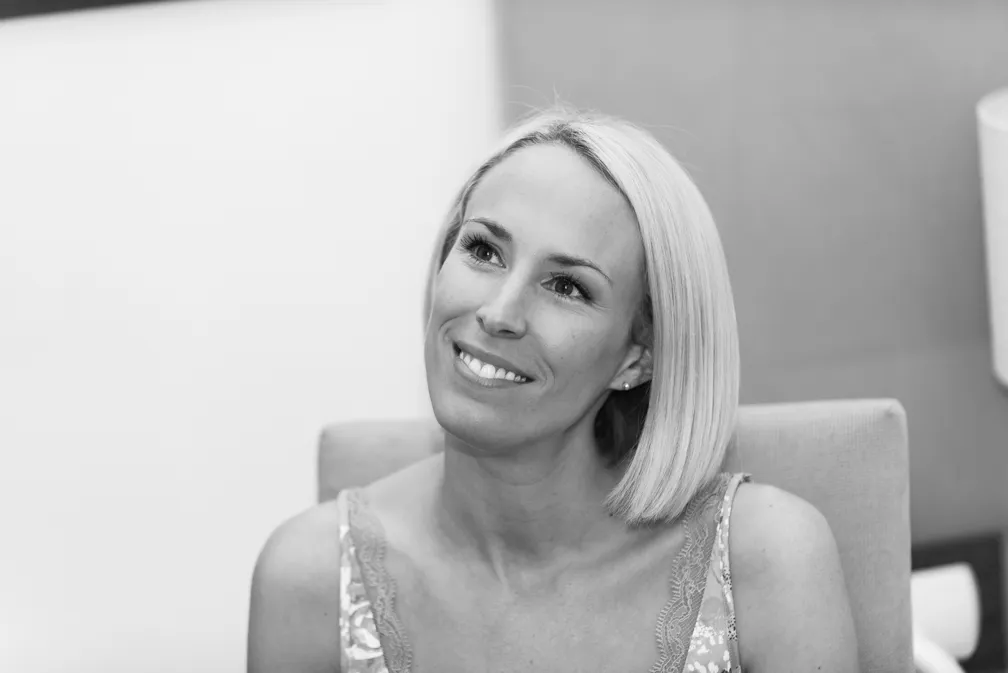 Smiling woman with straight blonde hair looking slightly upward while seated on a chair.
