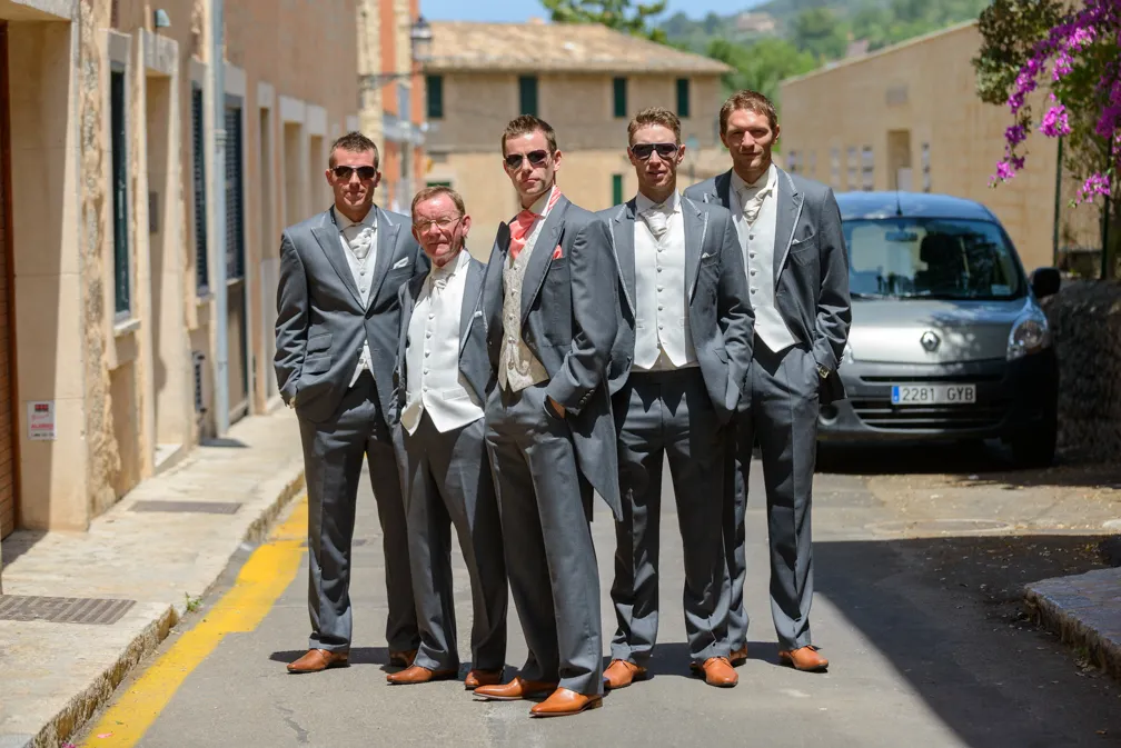 Five men in matching grey suits with white vests and brown shoes standing on a sunlit street in a Mediterranean village.