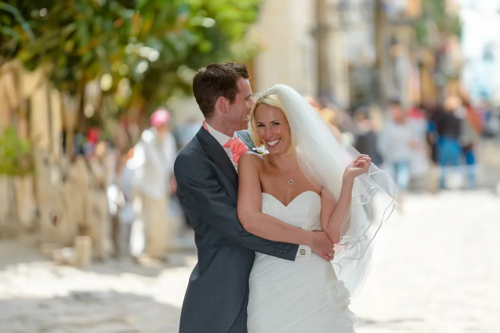 Bride in white strapless gown and veil smiling as groom in gray suit embraces her on sunlit street.