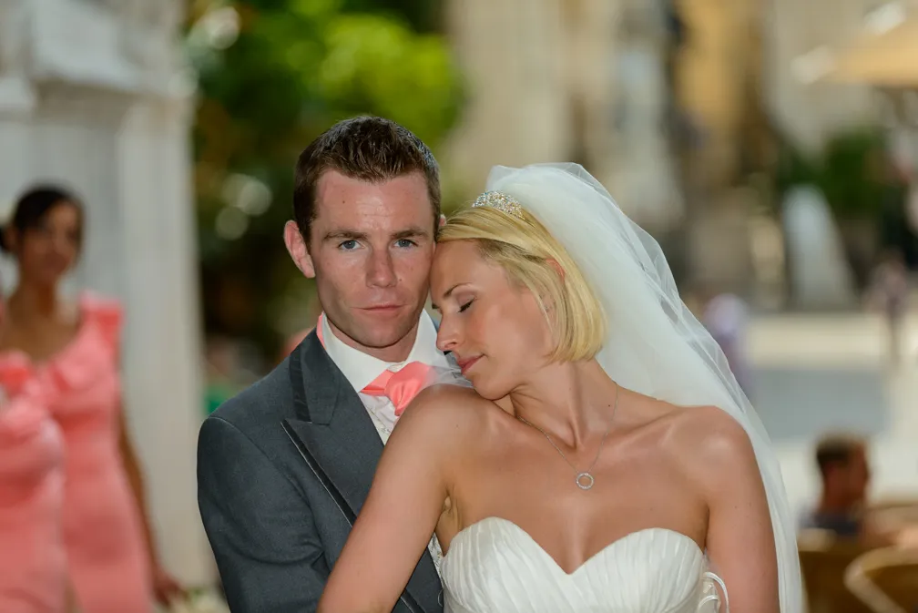 Bride leaning her head on groom's shoulder as he looks at the camera, both dressed in wedding attire with blurred guests in the background.