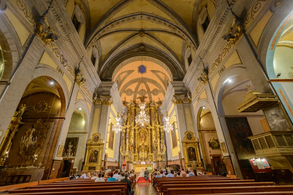 Interior of a historic church with ornate gold altar, chandeliers, vaulted ceilings, and people seated in wooden pews.