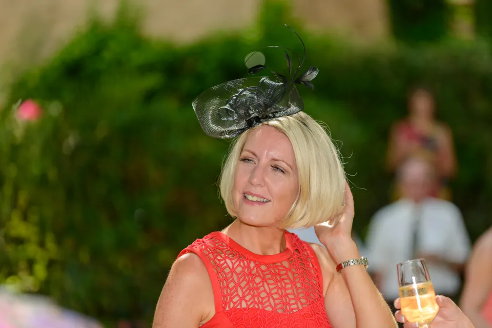 Blonde woman in a red lace dress wearing a black fascinator, holding her hair back outdoors at a social event.
