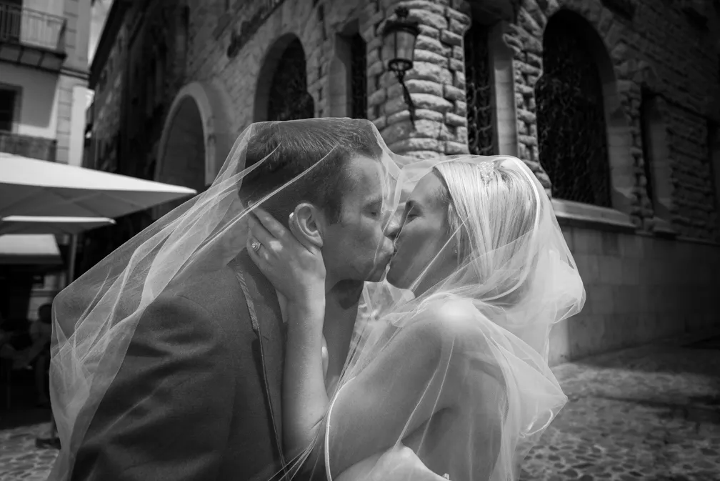 Black and white photo of a bride and groom kissing under the bride's veil in a cobblestone street with stone buildings in the background.