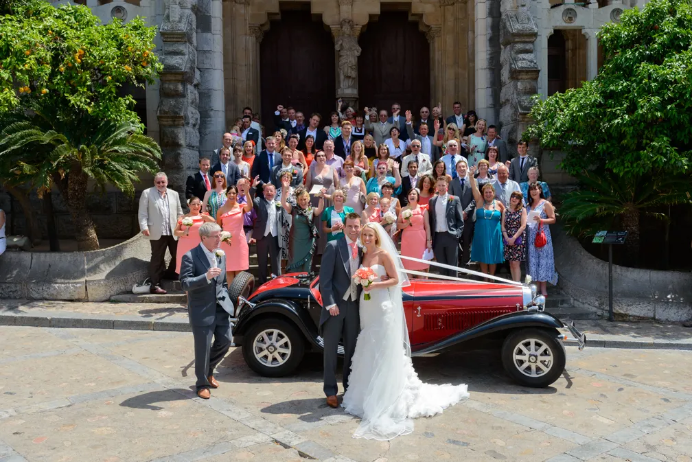 Bride and groom standing in front of a vintage red car with a large group of guests posing on stone steps outside a building.