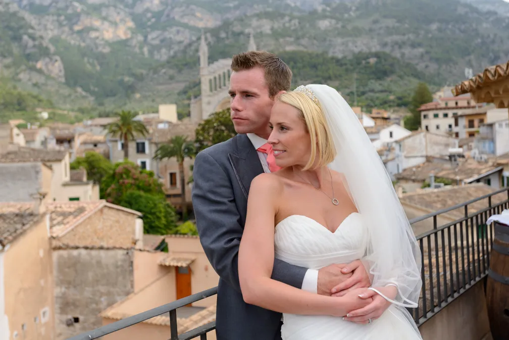 Bride in white wedding dress and veil embraced by groom in gray suit on balcony overlooking a village with mountains.