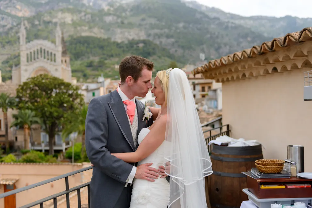 Bride and groom embracing on a balcony with mountainous village background.