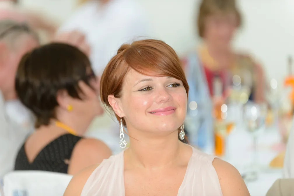 Smiling woman with red hair wearing dangling earrings at a social event with blurred guests and drinks in the background.
