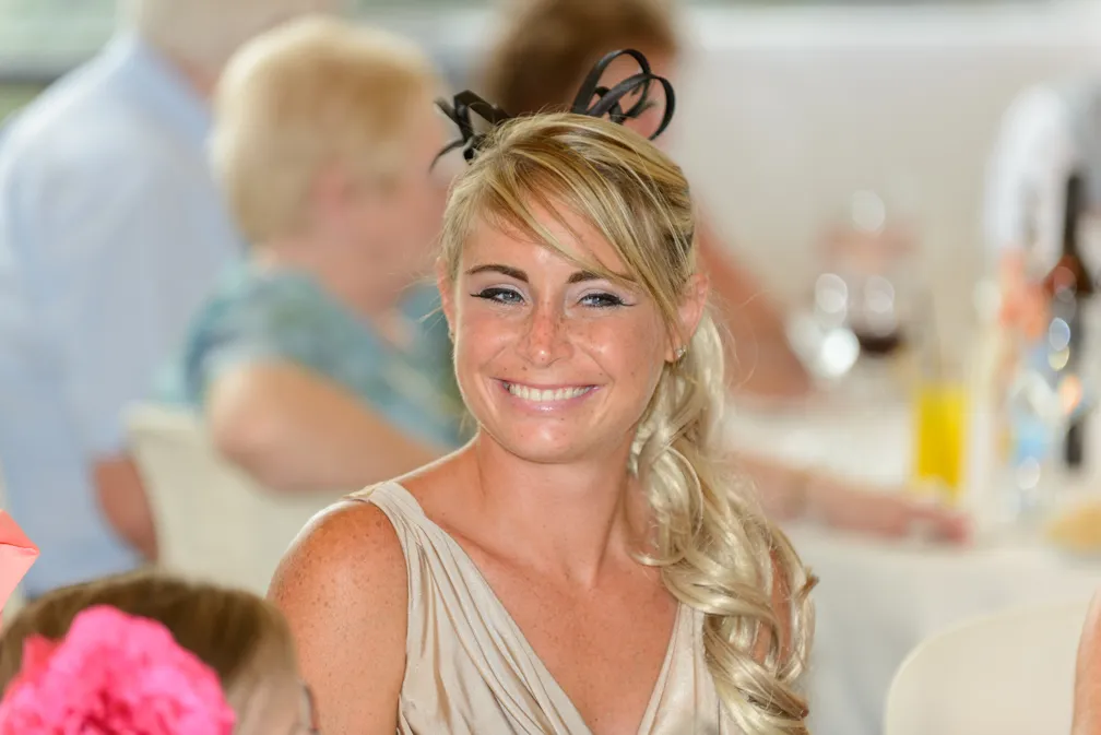 Smiling blonde woman with curled hair and black hair accessory at a social event with blurred guests and table in the background.