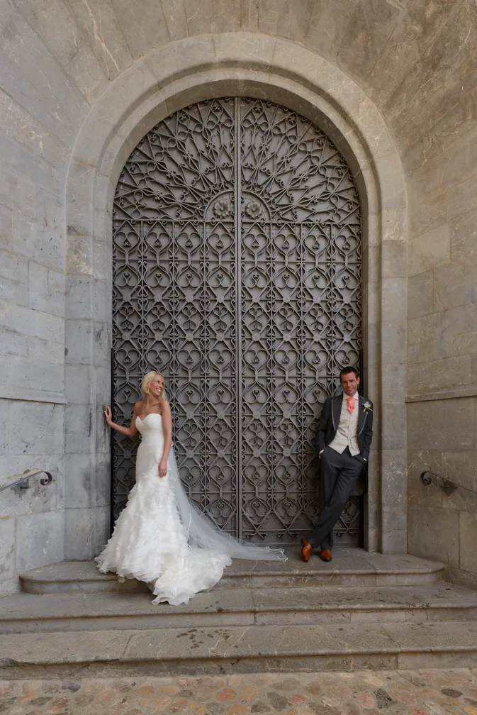Bride in white wedding dress and groom in suit posing in front of large ornate wrought iron door within stone archway.