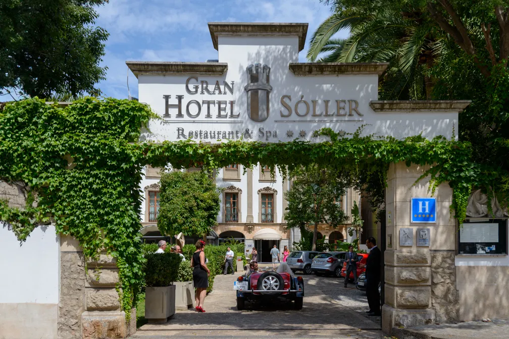 Entrance to Gran Hotel Sóller with green ivy-covered archway and people around vintage and modern cars.