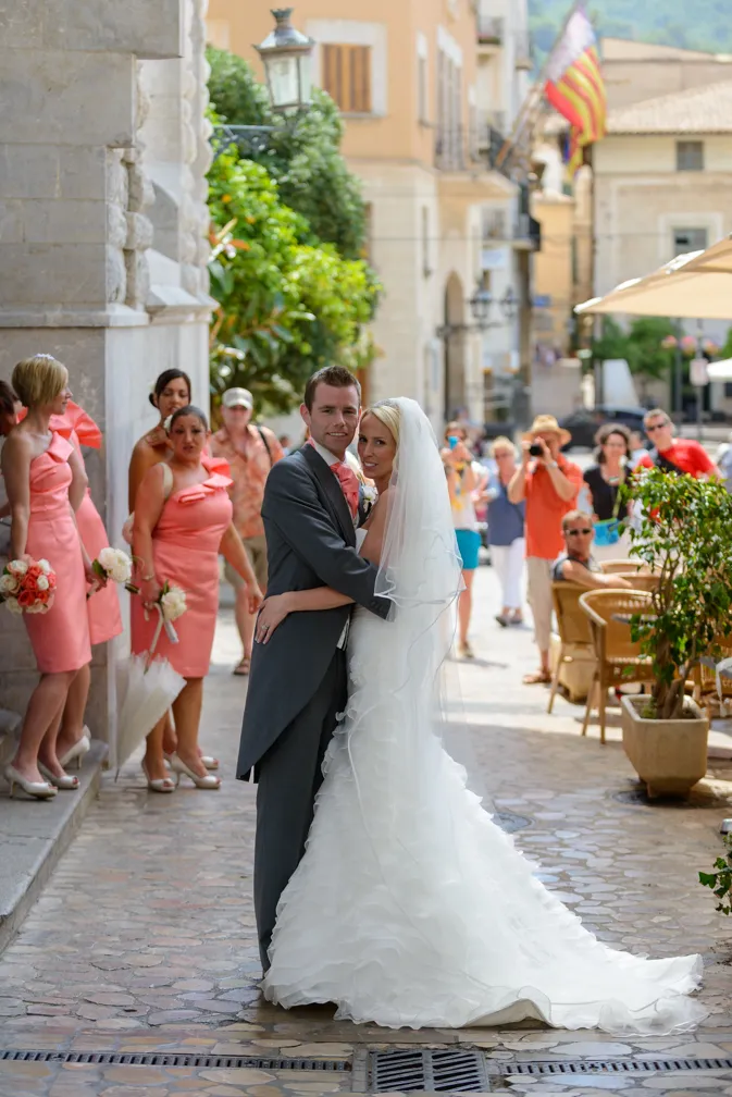Bride and groom embracing outdoors with bridesmaids in pink dresses and guests in the background.
