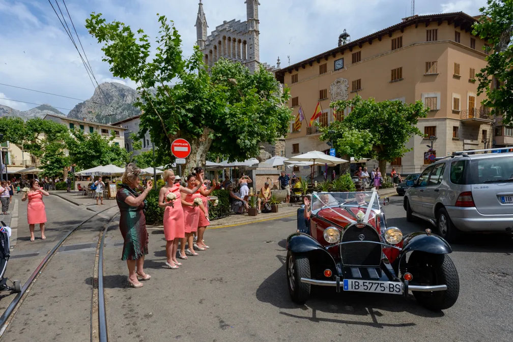 Vintage red and black convertible car driving on a street with women in coral dresses waving and taking photos, set in a sunny town square with stone buildings and trees.