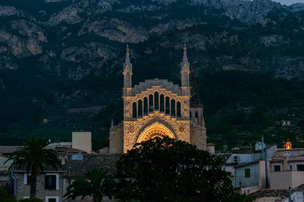 Illuminated stone church facade with tall arched windows and spires against a mountainous backdrop at dusk.