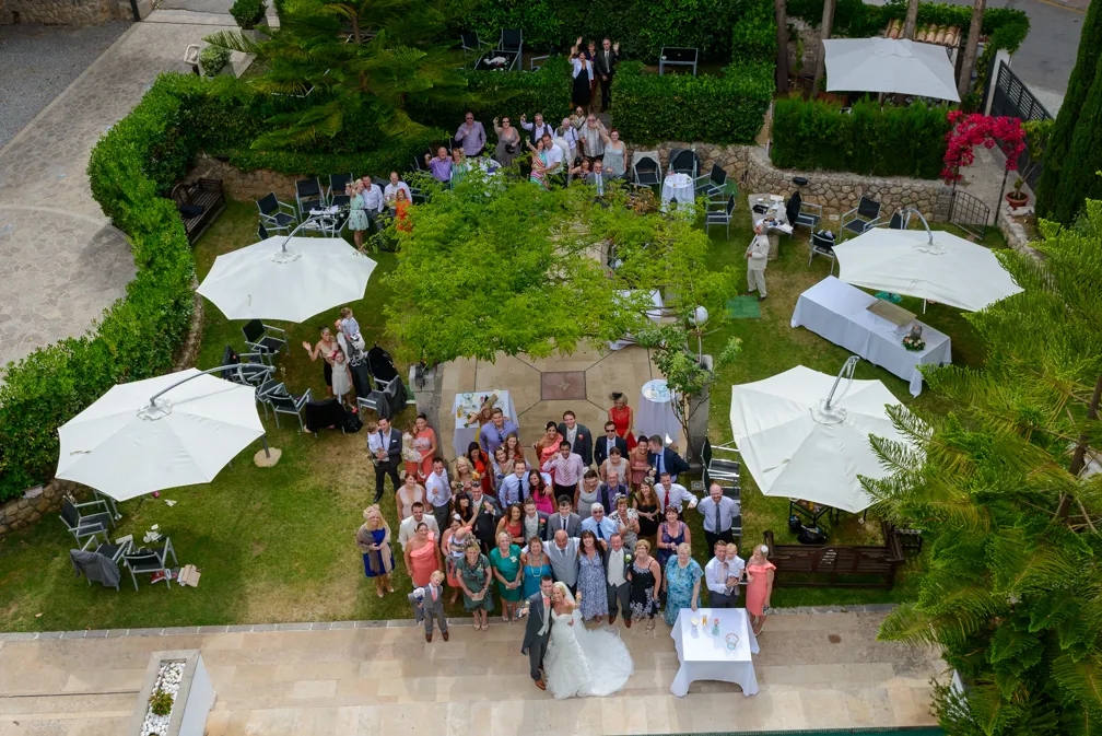 A large group of wedding guests posing around a bride and groom in an outdoor garden setting with white umbrellas and greenery.