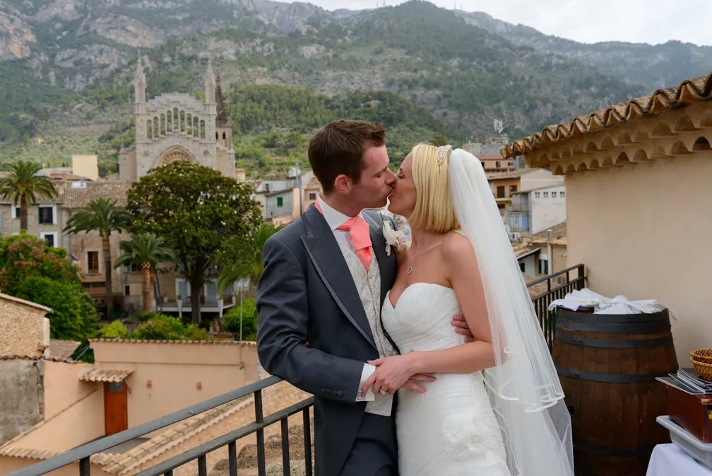 Bride and groom in wedding attire kissing on a balcony with a mountainous village and church in the background.