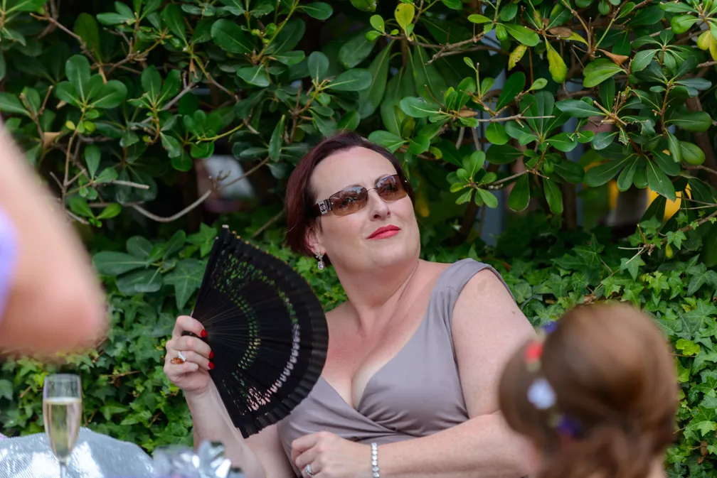 Woman wearing sunglasses and a gray sleeveless dress, fanning herself with a black folding fan while sitting outdoors near green bushes.
