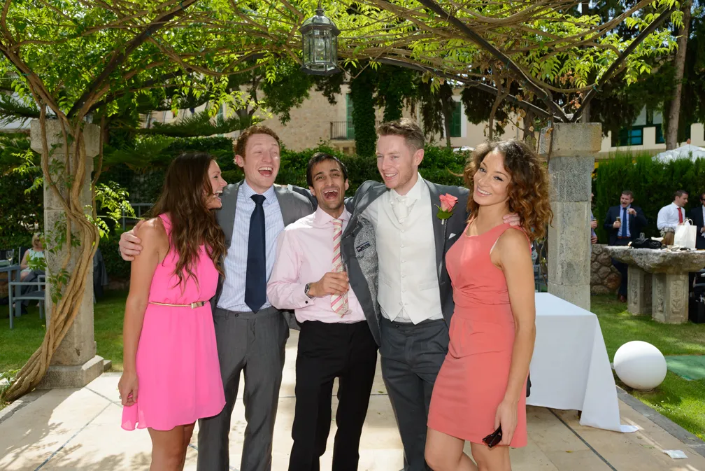 Five friends smiling and posing together outdoors under a vine-covered archway at a sunny garden event.
