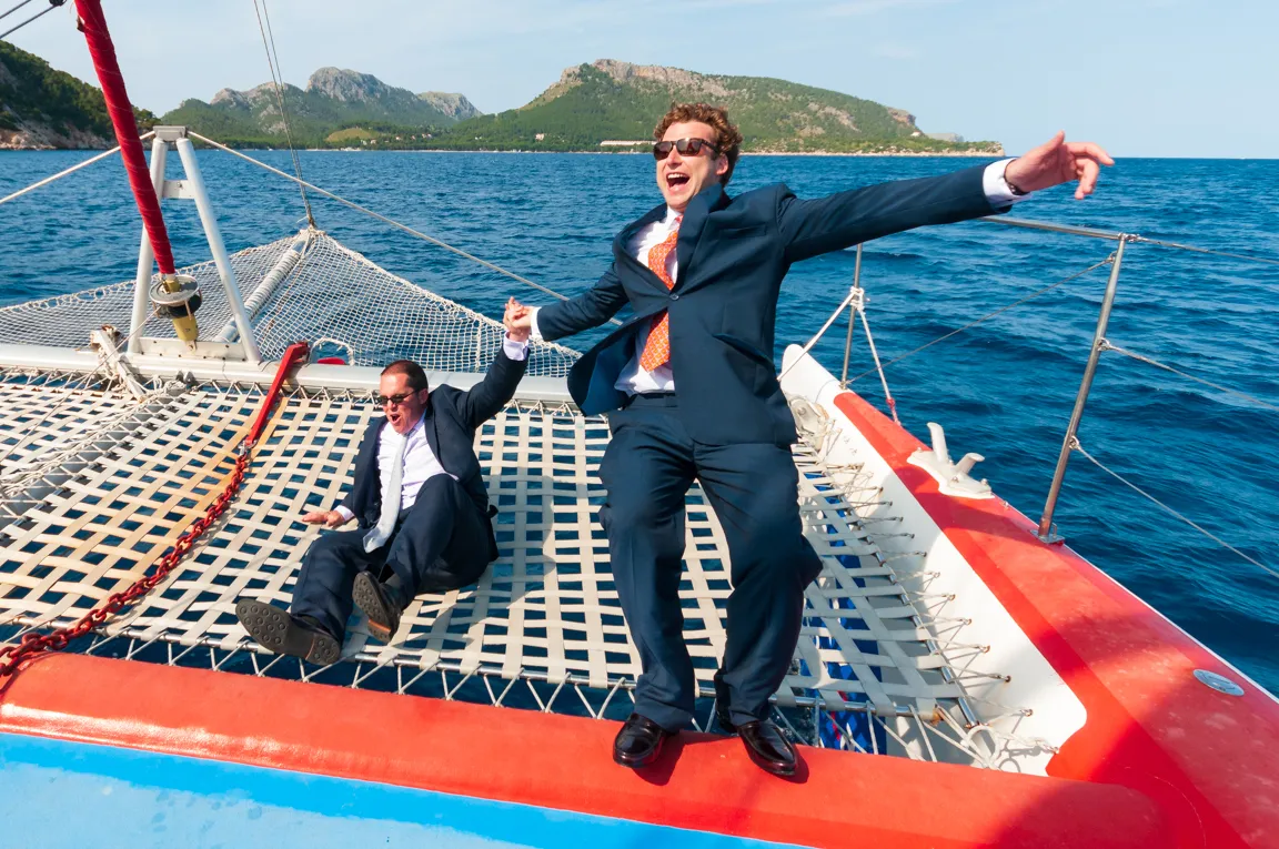 Two men in suits laughing and holding hands as they sit on the netting of a catamaran with sea and mountains in the background.