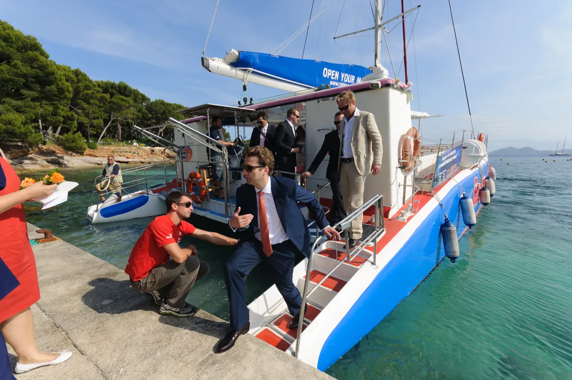 Men in suits boarding a blue and white sailboat docked at a seaside with clear water and pine trees in the background.