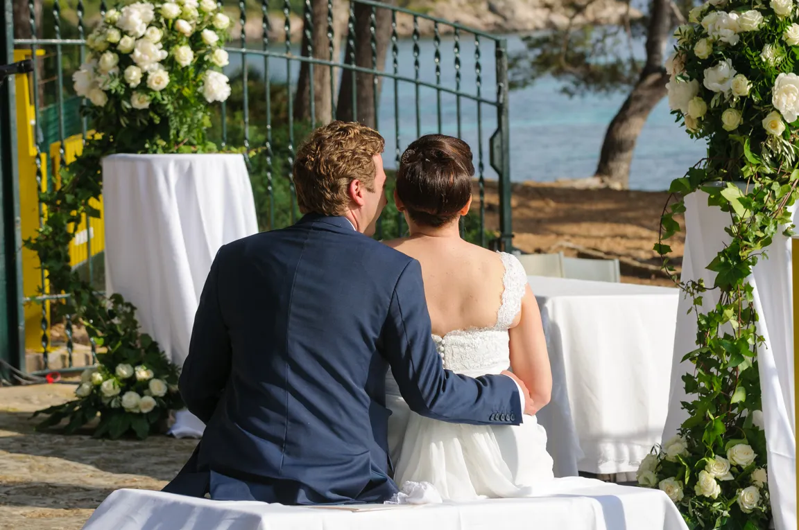 Bride in white lace dress and groom in navy suit sitting with their backs to the camera at an outdoor wedding ceremony with floral decorations.