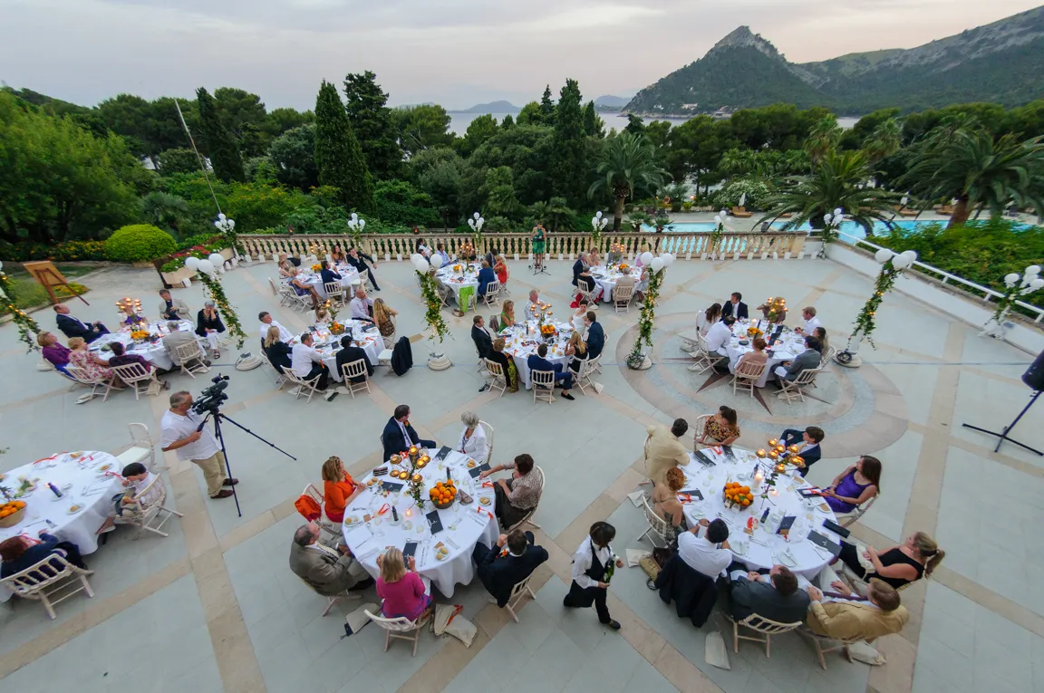 Outdoor formal dining event with multiple round tables set with white linens, guests seated, and a scenic mountain and sea background.