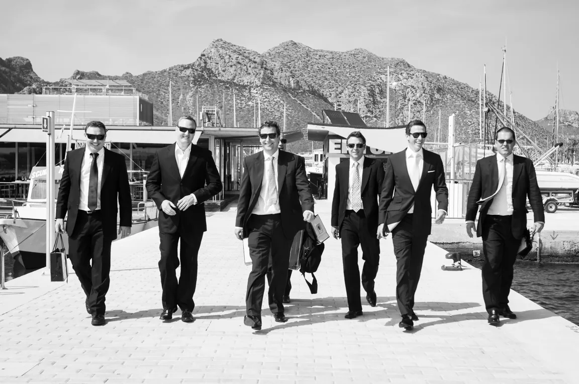 Six men in suits and sunglasses walking confidently on a dock with boats and mountains in the background.