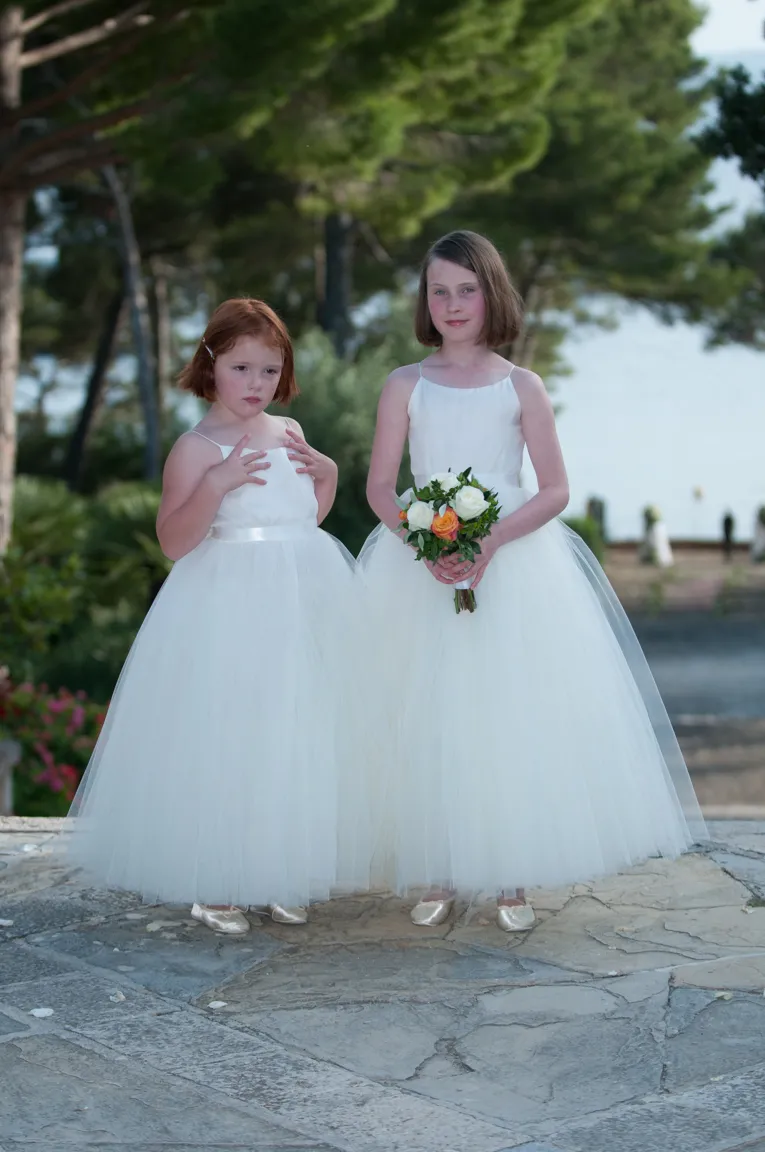 Two young girls in white tulle dresses, one holding a bouquet of flowers, standing outdoors on stone pavement with trees in the background.