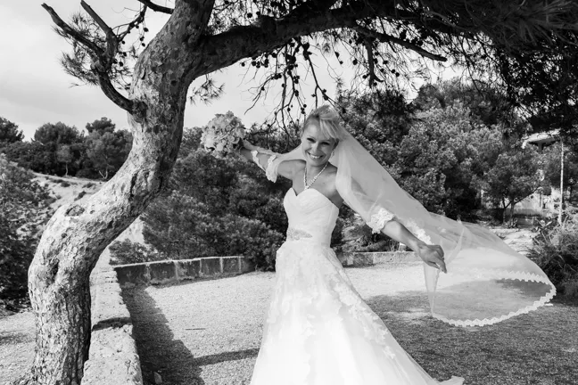 Smiling bride in a wedding gown holding bouquet and veil under a tree outdoors.
