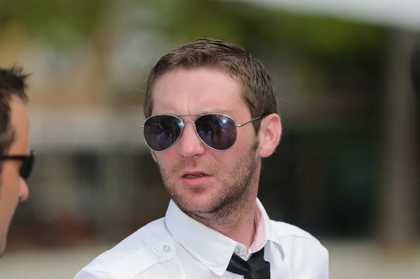 Man with short brown hair, wearing aviator sunglasses, a white shirt, and a loosened black tie, looking to the side.