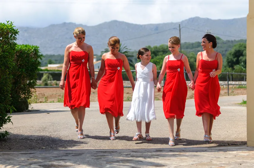 Four bridesmaids in matching red dresses holding hands with a young girl in a white dress, walking outdoors with mountains in the background.