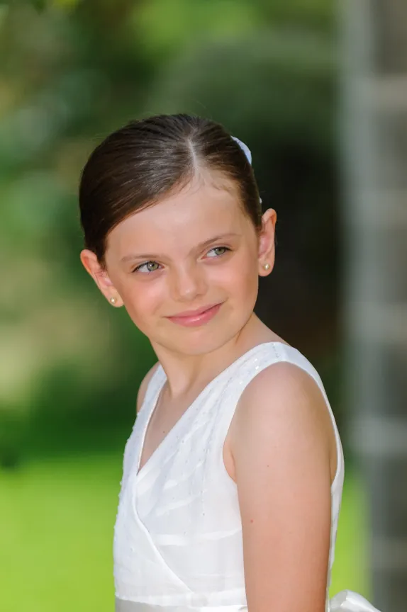Young girl with green eyes and dark hair in a white sleeveless dress smiling softly outdoors.