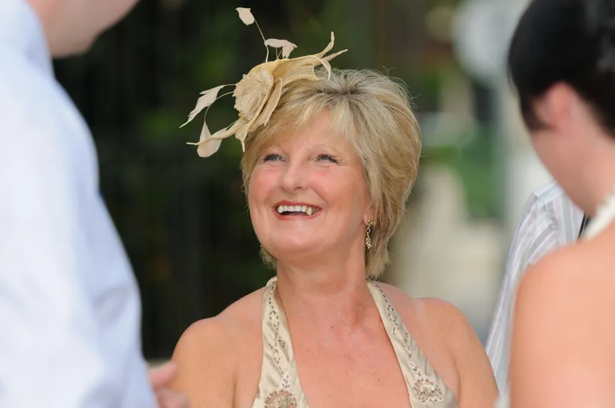 Smiling middle-aged woman wearing a beige dress and decorative fascinator at an outdoor event.