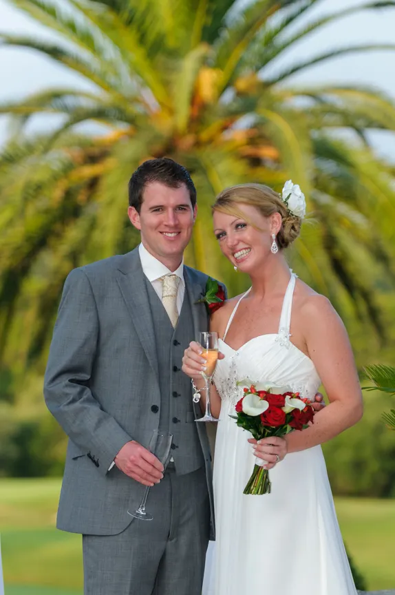 Bride in white dress holding a bouquet and groom in gray suit holding glasses, standing outdoors with palm tree in background.