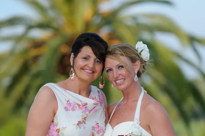 Two smiling women posing outdoors with a blurred palm tree background, one wearing a white floral dress and the other in a white halter dress with a flower in her hair.
