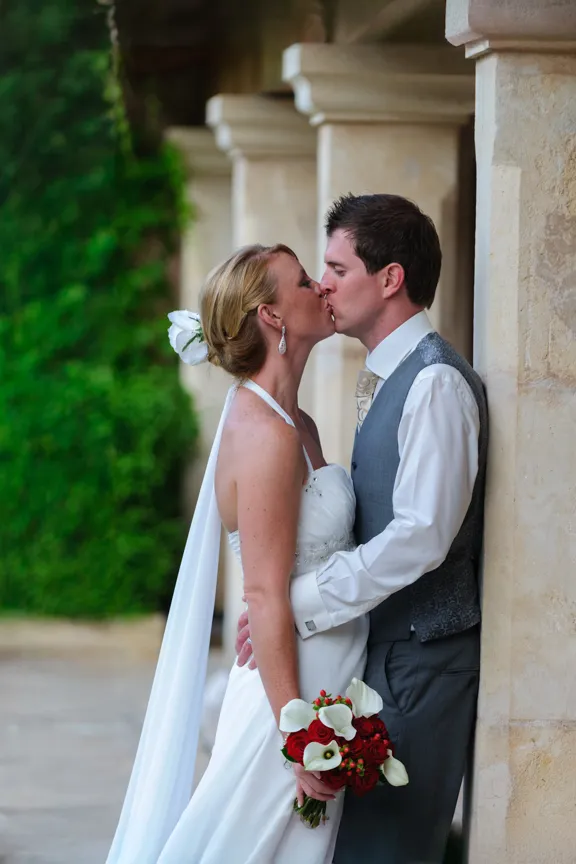 Bride and groom kissing by stone pillars, bride holding bouquet of red and white flowers.