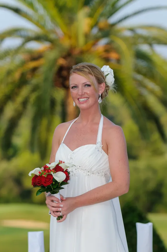 Smiling bride in a white halter wedding dress holding a bouquet of red roses and white flowers with a palm tree blurred in the background.