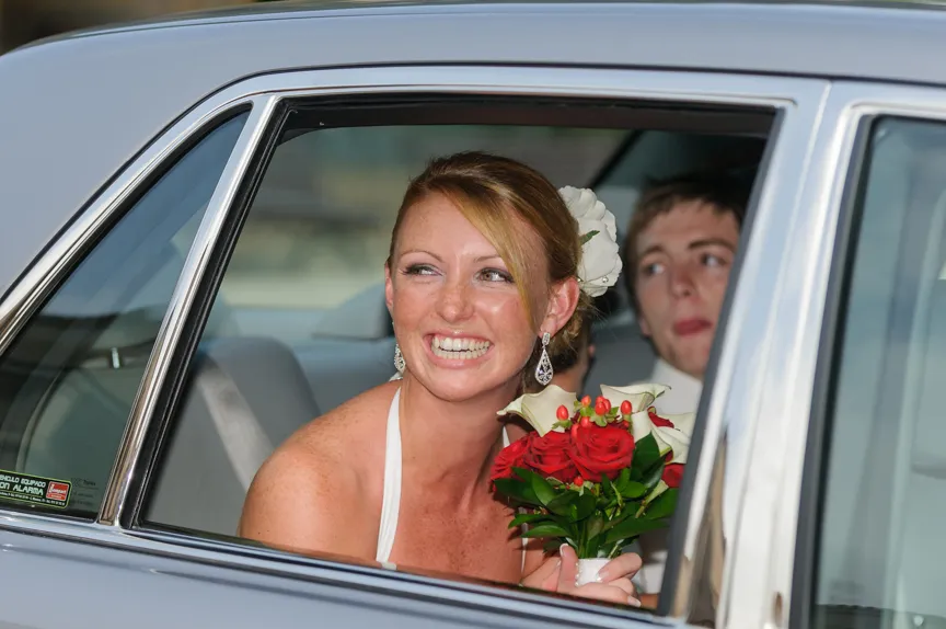 Bride smiling and holding a bouquet of red and white flowers sitting in a car with a groom in the backseat.